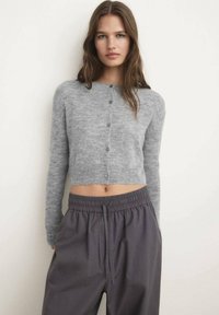 Lichtgrijze cropped cardigan met ronde hals, lange mouwen en vijf knopen. Gepaard met charcoal elastische taillebroek. Zachte textuur.