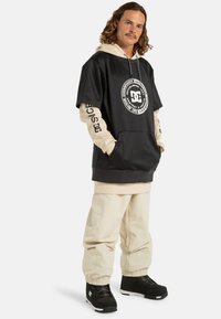 Zwarte oversized hoodie met wit cirkelvormig logo en beigekleurige lange mouwen, gedragen met beigekleurige losvallende broek en zwarte schoenen.