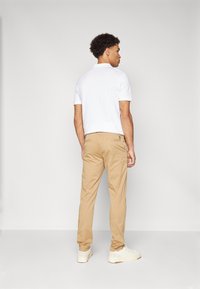Uomo in piedi di spalle, indossa una polo bianca a maniche corte, pantaloni beige e sneakers bianche con loghi sui talloni.