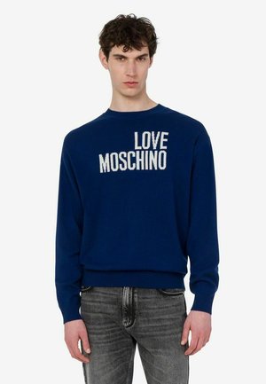 Młody mężczyzna w niebieskim swetrze z białym napisem "LOVE MOSCHINO" i wyblakłych szarych dżinsach stojący na jednolitym białym tle.