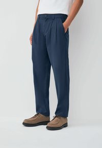 Person wearing navy blue pleated trousers with pockets and brown lace-up shoes, standing against a plain light background.