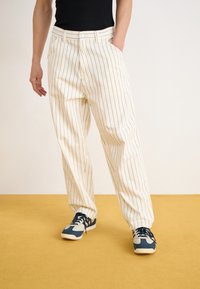 Weiß gestreifte Hosen mit lockerer Passform, ausgestattet mit Seitentaschen und einem Knopfverschluss, kombiniert mit blauen und grauen Sneakers.