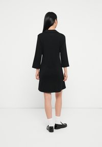 Czarna sukienka z kołnierzykiem, trzyczwartowym rękawem, dopasowanym topem i rozkloszowanym dołem, wykonana z gładkiego materiału. Noszona z czarnymi butami i białymi skarpetkami.