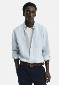 Camisa de botones a rayas azul claro con un bolsillo en el pecho, llevada abierta sobre una camiseta blanca. Combinada con pantalones oscuros y un cinturón marrón.
