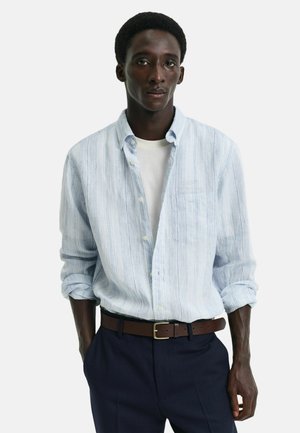 Camisa de botones a rayas azul claro con un bolsillo en el pecho, llevada abierta sobre una camiseta blanca. Combinada con pantalones oscuros y un cinturón marrón.