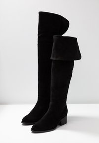 Bottes noires au-dessus du genou en daim, avec un revers plié en haut, un bout pointu et un talon bloc pour plus de stabilité.