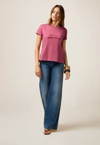Camiseta de algodón rosa de mangas cortas, con texto negro. Vaqueros de pierna ancha azules adornados con detalles de tachuelas plateadas a lo largo del costado.