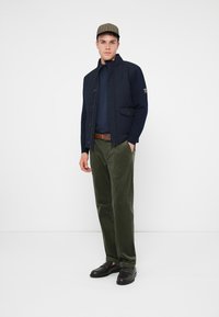 Marineblauwe gewatteerde jas met een hoge kraag en voorzakken, gedragen over een marineblauw topje, olijfgroene corduroy broek en zwarte instapschoenen.