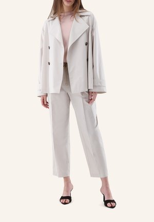 Frau trägt cremefarbenen, zweireihigen Blazer, passende hochgeschnittene Hose, hellrosa Oberteil und schwarze offene Sandaletten mit Absatz, stehend.