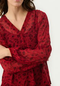 Blusa roja con estampado floral, escote en V y mangas largas elásticas. Hecha de una tela translúcida, cuenta con un diseño texturizado y un corte fluido.