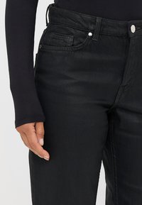 Jeans de mezclilla negra de cintura alta, con bolsillos delanteros, herrajes plateados y una textura suave. Mano apoyada en el bolsillo lateral.