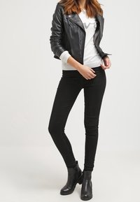 Vrouw poseert in een zwart leren jack, witte trui, zwarte skinny jeans en zwarte enkellaarsjes tegen een effen lichte achtergrond.