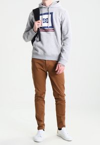 Grå huvtröja med en grafisk logotyp på framsidan, kombinerad med bruna byxor och vita sneakers. Modellen håller en svart ryggsäck.