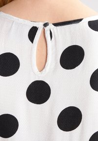 Chemisier blanc à pois noirs ; présente une ouverture en trou de serrure à l'arrière, une texture douce et une fermeture à bouton au niveau du cou.