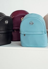 Light blue faux leather backpack with a rounded top, front zipper pocket, and metallic logo accent. Other bags in black, burgundy, and beige.