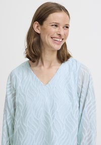 Blusa azzurro chiaro con scollatura a V e ricamo a motivo ondulato. Le maniche lunghe semi-trasparenti aggiungono texture e profondità al design.