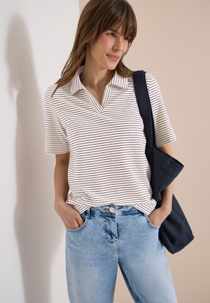 Femme aux cheveux bruns porte un haut à manches courtes rayé blanc et rouge, un jean bleu clair et porte un sac fourre-tout bleu foncé sur l'épaule.