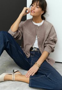 Beige suedine jas met drukknoopjes, gedragen over een witte blouse; gecombineerd met donkerblauwe wijde jeans en beige block-heeled schoenen. Accentuering met een gouden armband.