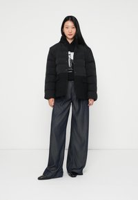 Melnā pūkainā jaka ar teksturētu apakšējo paneli, pogu aizdari un kapuci, savienota ar brīviem tumšiem džinsiem un melnām kurpēm.