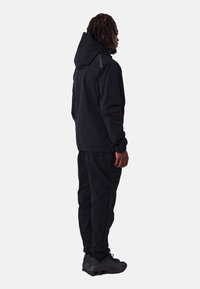 Chaqueta cortavientos negra con capucha, puños elásticos y ligero brillo, combinada con pantalones joggers negros a juego y zapatillas oscuras.