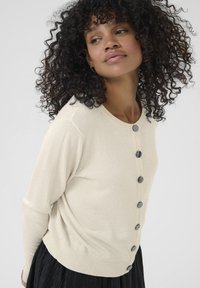 Woman with curly dark hair wearing a cream buttoned cardigan and black skirt, looking slightly to the side against a plain background.