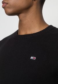 Svart stickad tröja med rund halsringning och ribbad krage. Har en liten broderad logotyp i röd, vit och blå på bröstet.