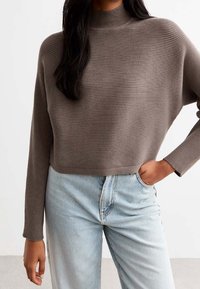 Brown ribbed knit sweater with a high neckline, long sleeves, and a cropped cut, paired with light blue high-waisted jeans.
