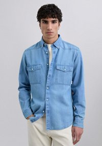 Camisa de mezclilla azul claro de manga larga, con cierre de botones en la parte delantera, dos bolsillos en el pecho y una textura suave. Combinada sobre una camisa blanca.
