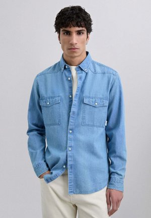 Camisa de mezclilla azul claro de manga larga, con cierre de botones en la parte delantera, dos bolsillos en el pecho y una textura suave. Combinada sobre una camisa blanca.
