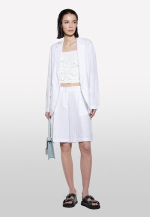 Blazer en lin blanc sur un haut court blanc à texture avec des accents floraux. Shorts plissés blancs et sandales noires avec des détails en métal argenté. Sac bleu clair.