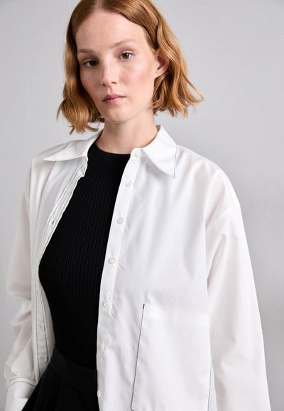 Cămașă albă cu guler ascuțit, mâneci lungi și cusături în contrast. Purtați-o peste un top negru cu dungi, punând în evidență o siluetă elegantă.