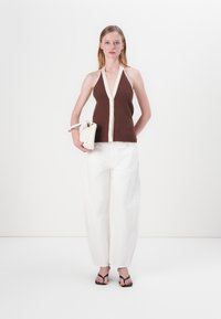 Hnedý rebrovaný halter top s krémovými gombíkmi a lemovaním, skombinovaný s bielymi širokými nohavicami a čiernymi sandálmi na remienky. Biela kabelka s detailom.