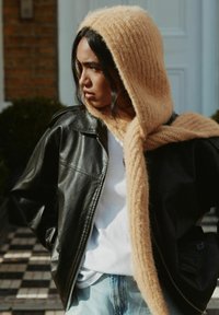 Schwarze Lederjacke, crèmefarbener Strickschal mit gerippter Textur, weißes Oberteil und hellblaue Jeans. Außenszene mit einem gemusterten Stein-Hintergrund.