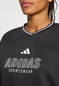 Kamera je fokusirana na osobu koja nosi crnu Adidas sportsku majicu s bijelim logotipom i kontrastnim ovratnikom. Pozadina je neutralna.