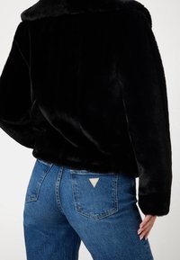 Schwarze Jacke aus Kunstpelz mit weitem Kragen, lockerem Schnitt und langen Ärmeln, kombiniert mit hoch taillierten blauen Denim-Jeans mit einem dreieckigen Logo.