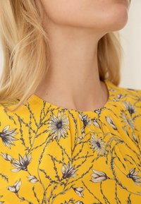 Femme blonde portant un chemisier jaune avec un motif floral noir et blanc, montrant le décolleté et une partie du visage tournée vers le haut.