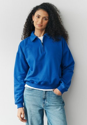Frau mit lockigem schwarzen Haar, die einen blauen Pullover mit einem halben Reißverschluss und hellblauen Jeans trägt, steht vor einem weißen Hintergrund.