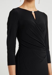 Czarna sukienka z rękawami o długości trzy czwarte, z dekoltem w kształcie klucza i marszczoną strukturą w talii. Gładki, elastyczny materiał z błyszczącym akcentem.