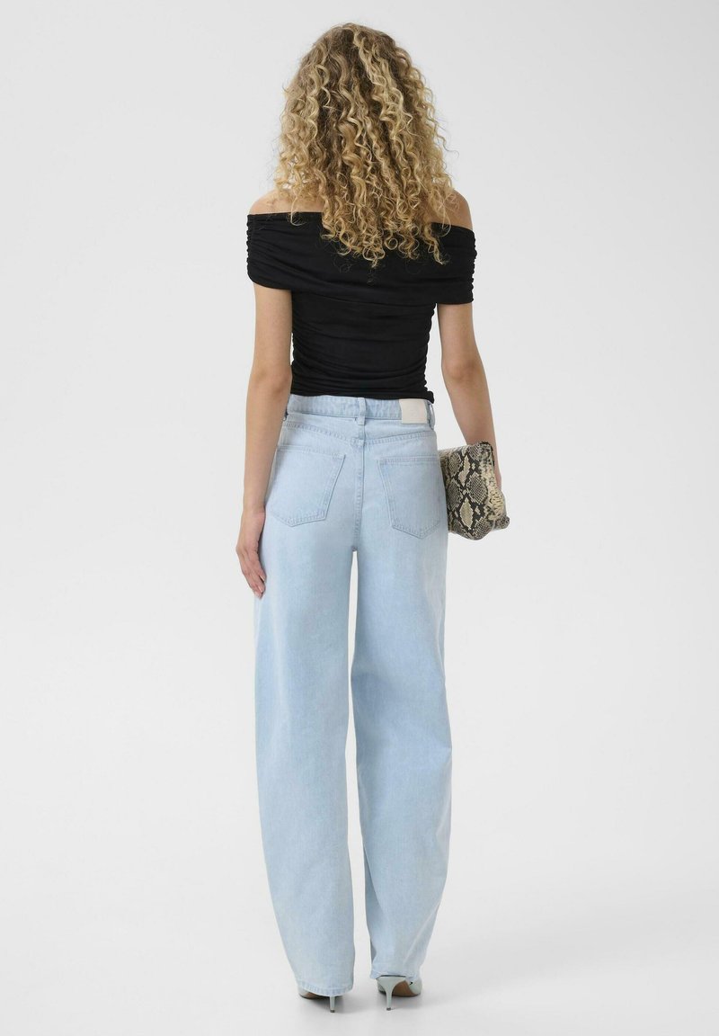 Vrouw met krullend haar, gekleed in een zwarte off-shoulder top, lichtblauwe wijd uitlopende jeans, met een clutch van slangenleer, staand met haar rug naar de camera.