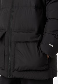 Chaqueta acolchada negra con cremallera frontal, dos grandes bolsillos con solapa y puños elásticos. El material parece aislante y suave.