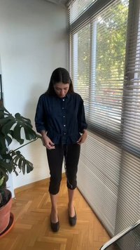 Γυναίκα στέκεται μέσα κοντά σε μεγάλες περσίδες παραθύρου και ρυθμίζει το κορδόνι του σκούρου μπλε πουκάμισού της με κουμπιά, με μια γλάστρα κοντά της.