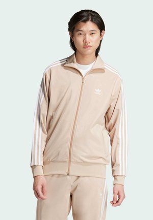 Beige zip-up träningsjacka i slät tyg, med vit tredelad design på ärmarna och en liten vit logotyp på bröstet.