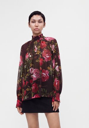 Mujer con cabello corto y oscuro que lleva una blusa transparente oscura con flores y mangas largas y una falda negra contra un fondo liso y claro.