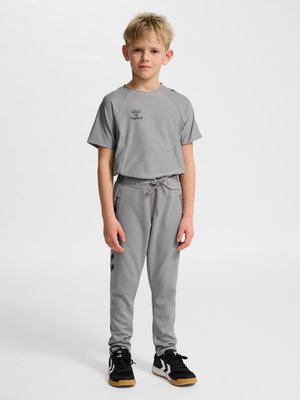 Jeune garçon debout, face à l'avant, portant des vêtements de sport gris avec des logos et des baskets noires avec des accents blancs.