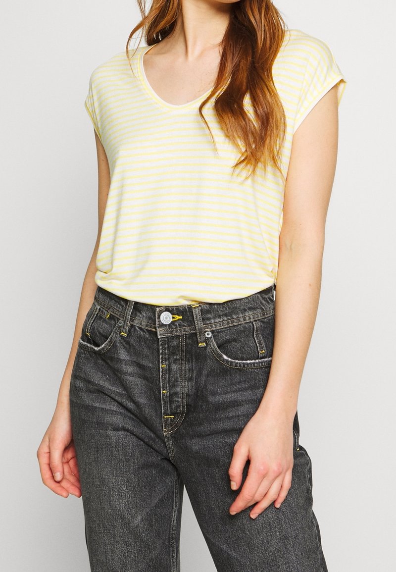 Žluté a bílé pruhované tričko s kulatým výstřihem, v kombinaci s černými džíny s vysokým pasem, které mají žluté švy a klasický střih.
