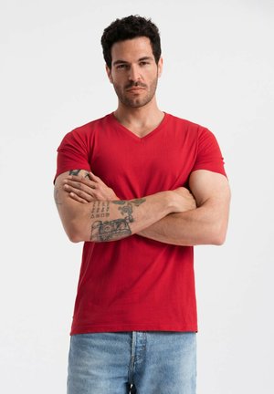 Rød bomulds T-shirt med V-hals design, korte ærmer og en afslappet pasform. Viser synlige tatoveringer på underarmen.