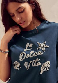 Tumedate sinine sweat-jakk, millel on tekstuuriga ja kristallidega kaunistatud disain, millel on fraas "La Dolce Vita" ning tähtede ja kilpkonnade motiivid.