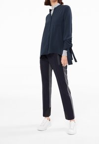 Marinblå långärmad skjorta med knappar, bröstficka och snörning på ärmarna, tillsammans med mörka byxor med olika tygtexturer och vita sneakers.