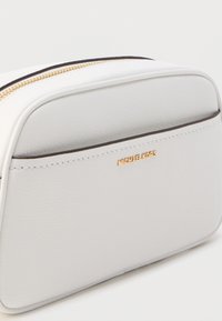 Valkoinen nahkainen kosmetiikkalaukku kullanvärisillä yksityiskohdilla, pyöreä muoto, vetoketjusuljenta, etupuolella pieni logo ja kontrastiväriset tummat reunukset.