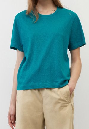 Osoba w turkusowej koszulce z krótkim rękawem i beżowych spodniach, stojąca z jedną ręką w kieszeni na jednolitym tle.